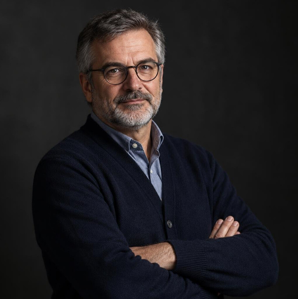 Studio portrait of a middle-aged Croatian male mystery writer with grey beard, glasses and a dark cardigan, arms crossed, neutral dark background